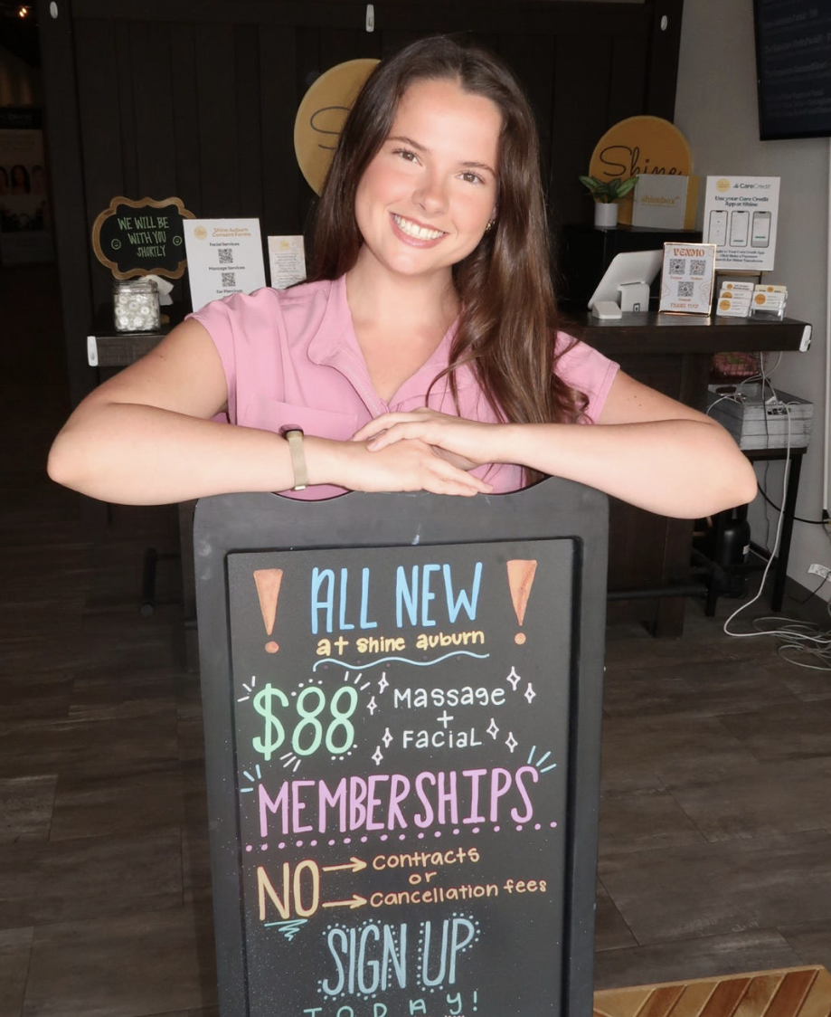A Shine Auburn esthetician standing behind a chalkboard sign announcing $88 massage and facial memberships